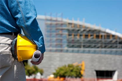 A full shot of a construction worker holding a yellow hard hat and a roll of paper on his side against a blurry building background.